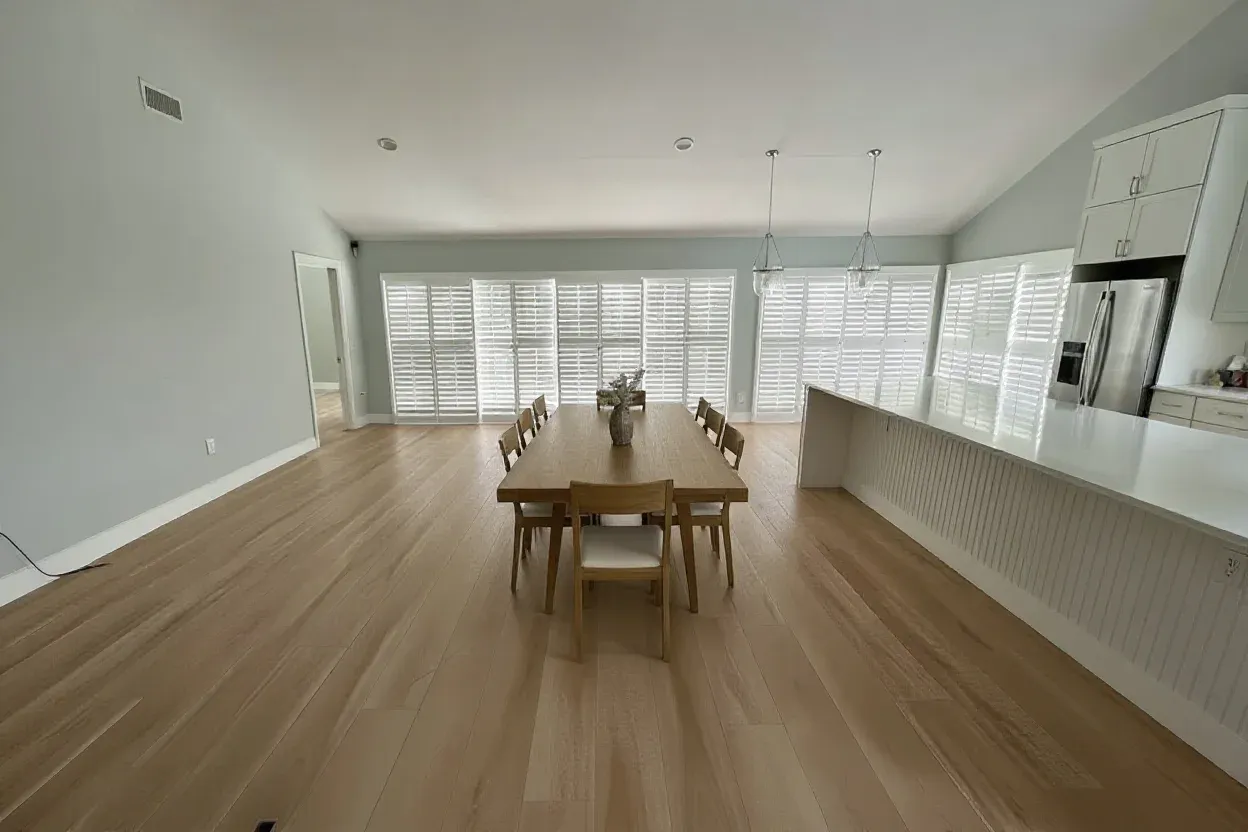 Spacious, minimalist dining area with a light wood table, eight chairs, and a vase centerpiece. Sunlight streams through large shuttered windows.